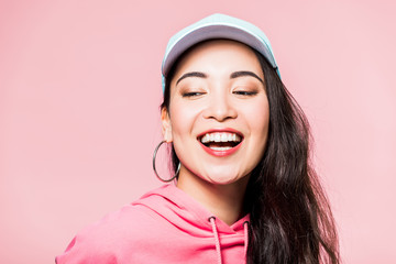 attractive asian woman in pink pullover and cap smiling isolated on pink