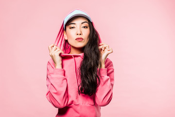 Naklejka premium attractive asian woman in pink pullover and cap looking at camera isolated on pink