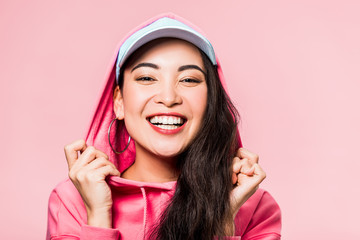 attractive asian woman in pink pullover and cap smiling isolated on pink