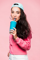 attractive asian woman in pullover and cap drinking from plastic cup isolated on pink