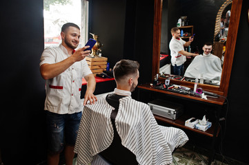 Young bearded man getting haircut by hairdresser while sitting in chair at barbershop. Barber soul. Master making photo on phone.