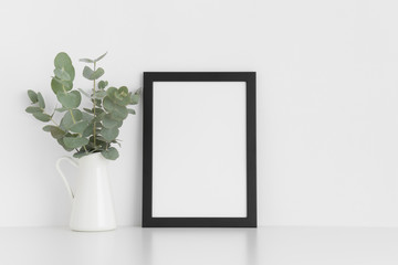 Black frame mockup with a bouquet of eucalyptus in a vase on a white table.Portrait orientation.