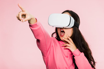 shocked woman in pink pullover with virtual reality headset pointing with finger isolated on pink