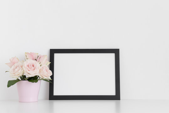 Black frame mockup with pink roses in a pot on a white table.Landscape orientation.