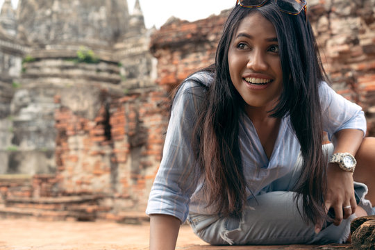 Attractive happy Asian girl outdoors sitting on brick wall looking to the side - Local tour guide woman exploring ancient cultural relics on day trip to historical park - tourism and activity concept