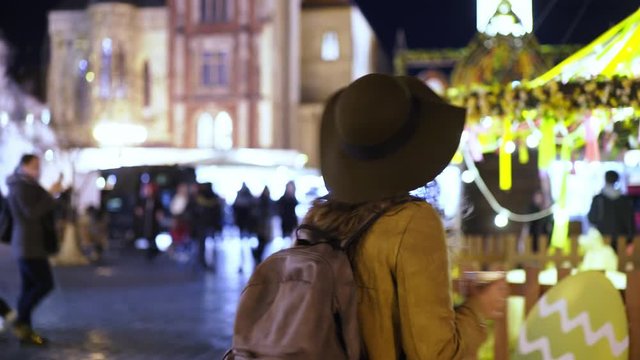 Elegant woman in hat walking on traditional easter fair in downtown of Prague at night. Medium shot young girl drinking mulled wine and goes toward the camera through decorated and illuminated market