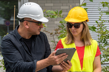 Two workers on construction site wearing safety vest and helmet checking the work plan on a digital tablet computer