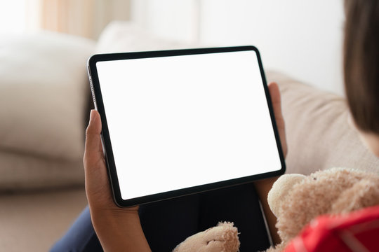 Mockup, Blank digital tablet in the hand of Asian  little girl, Tablet  white screen. While sitting on the sofa at her home. 