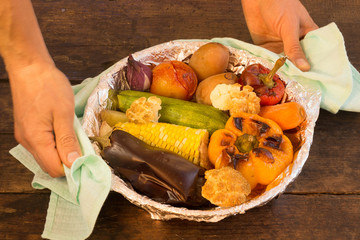 Male hand holding baked vegetables in foil potato carrot pepper onion corn on the cob eggplant cauliflower squash on wooden table