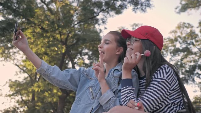 Cute cheerful girls friends gesturing and grimacing while making selfie photo on smartphone in the park