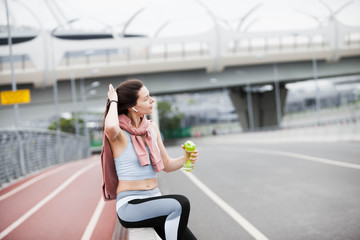 Fototapeta na wymiar Athletic woman in sports outfit after jogging, drinks water from bright green bottle, sitting on the parapet of bridge.
