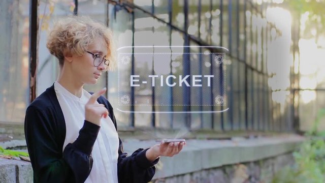 Curly young woman in glasses interacts with a hud hologram with text E-ticket. Blonde girl in white and black clothes uses technology of the future mobile screen