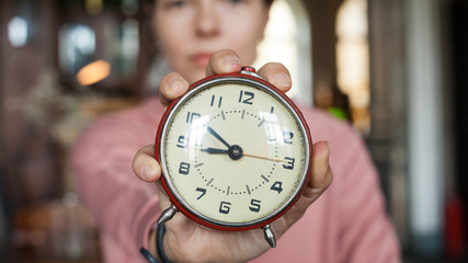 Deadline. A closeup alarm clock aimed at the frame held by a brunette girl in a pink sweater at...