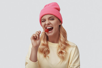 Attractive young woman smiling and eating lollipop while standing against grey background