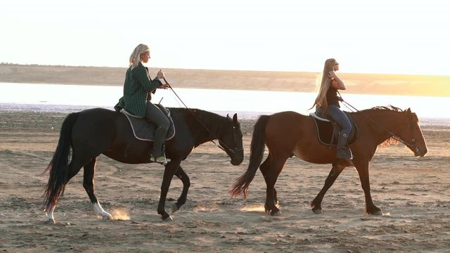 View of women riding horses along the river in golden light sunset or sunrise. Stallion walking in desert by the water. Slow motion