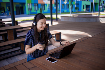 Girl with long hair sits at wooden table outdoor, looks at laptop monitor, drinks coffee, laughs and talks over the net.