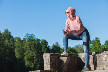 Young woman wearing sunglasses and hood on his head holding a remote control, carefully looks into the distance against.
