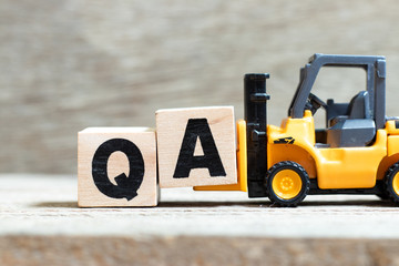 Toy forklift hold letter block a to complete word (Abbreviation of Quality Assurance or question and answer)on wood background