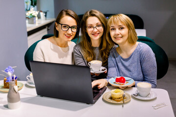 Women friends looking at camera in cafe, eating tasty desserts and drinking tea, using laptop for work. Women bloggers freelancers working in cafe