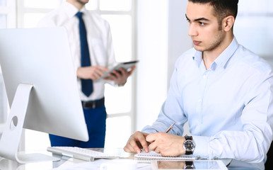 Businessman working with computer in modern office, male colleague at background . Headshot of arab male entrepreneur or manager at workplace. Business concept, teamwork and multi ethnic