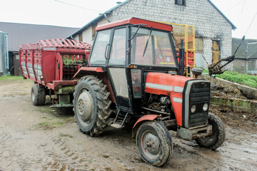 Obraz premium Red tractor with a trailer in the yard of a dairy farm. Side view. A barn in the background. Podlasie, Poland.