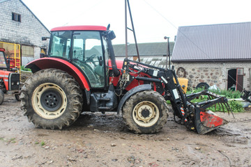 Red tractor with a lift for various works in the yard of a dairy farm. Side view. A barn in the background. Podlasie, Poland.