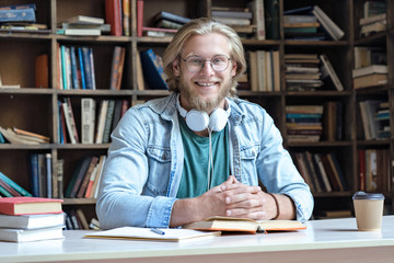 Happy guy university student in library learning with books headphones doing work assignment looking at camera sit at desk, smiling young man teacher wear glasses study in campus, portrait