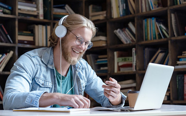 Happy male student online teacher laugh wear headphone talk video calling on laptop computer having fun communicate with skype tutor concept, distance education e learning chat course in library
