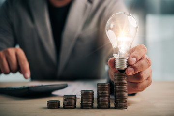 businessman hand holding lightbulb with coins stacking on desk.saving energy and money concept.