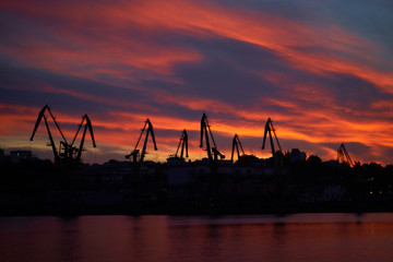 Obraz premium Colorful sunset over sea port and industrial cranes. Bright sunset in seaport. Large silhouettes of cargo cranes. Beautiful landscape with fiery sunset sky and sea.