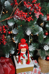 A cute children's soft doll in a red hat stands next to a Christmas tree. New Year's gift children's soft doll on the background of the New Year tree