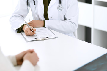 Woman doctor and patient sitting and talking at medical examination at hospital office, close-up. Therapist filling up medication history records. Medicine and healthcare concept