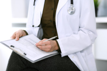 Woman doctor writing something at clipboard while sitting at the chair, close-up. Therapist at work filling up medication history records. Medicine and healthcare concept