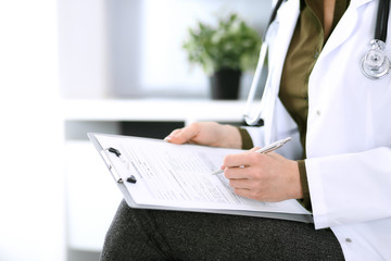 Woman doctor writing something at clipboard while sitting at the chair, close-up. Therapist at work filling up medication history records. Medicine and healthcare concept