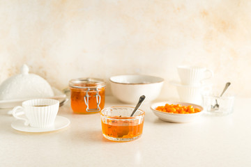 Buck seathorn jam in glass jars, white tea cups with saucers, bowls all set for breakfast with pastel colored table and wall
