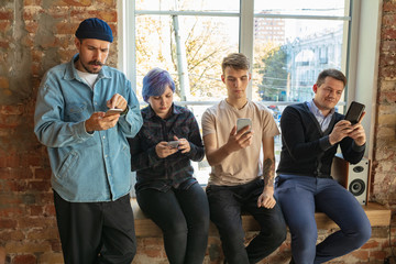 Group of happy caucasian young people standing behind the window. Sharing a news, photos or videos from smartphones, talking or playing games and having fun. Social media, modern technologies.