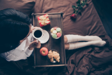 Cropped top above high angle view of nice attractive girl sitting on bed long legs wearing gaiters drinking coffee eating fresh delicious homemade baked snack on sheets linen house flat