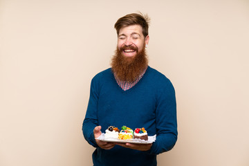 Redhead man with long beard over isolated background holding mini cakes