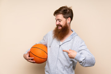Redhead man with long beard over isolated background with ball of basketball and pointing it
