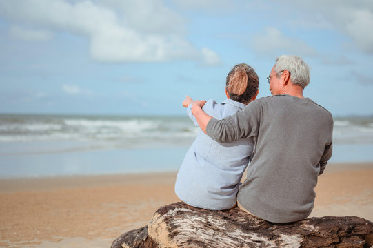 A Retired Couple Sitting At The Sea, A Woman Pointing A Finger For A Man To Watch Something On The Rocks And Holding Hands At The Beach In The Morning, Life Insurance Plans At Retirement Concepts.