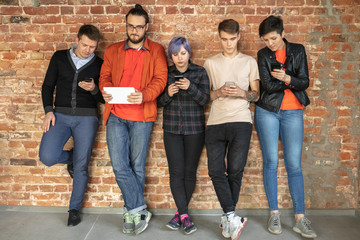 Group of happy caucasian young people standing behind the brick wall. Sharing a news, photos or videos from smartphones or tablets, playing games and having fun. Social media, modern technologies.