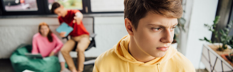panoramic shot of pensive boy in yellow hoodie and laughing classmates bullying him