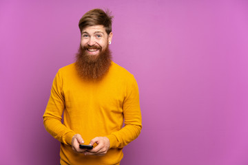 Redhead man with long beard over isolated purple background sending a message with the mobile