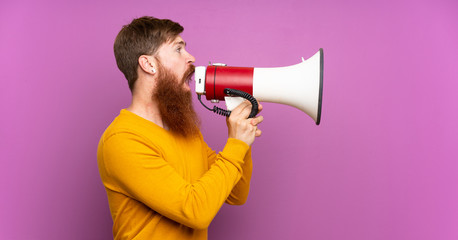 Redhead man with long beard over isolated purple background shouting through a megaphone