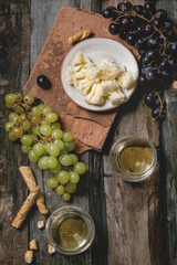 Swiss cheese tete de moine, black and green grapes and two glasses of white wine, served with bread sticks on old wooden plank background. Flat lay, space