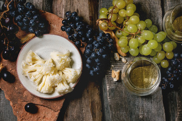 Swiss cheese tete de moine, black and green grapes and two glasses of white wine, served with bread sticks on old wooden plank background. Flat lay, space