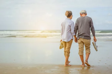 Fotobehang Senior couple walking on the beach holding hands at sunrise, plan life insurance at retirement concept. © Day Of Victory Stu.