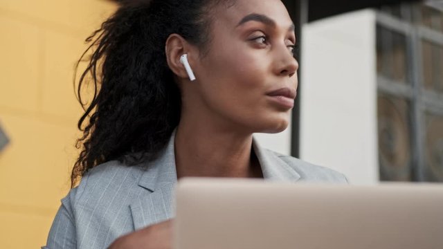 Close up view of serious young african business woman listening radio with earpods and waiting for someone while working on laptop in cafe