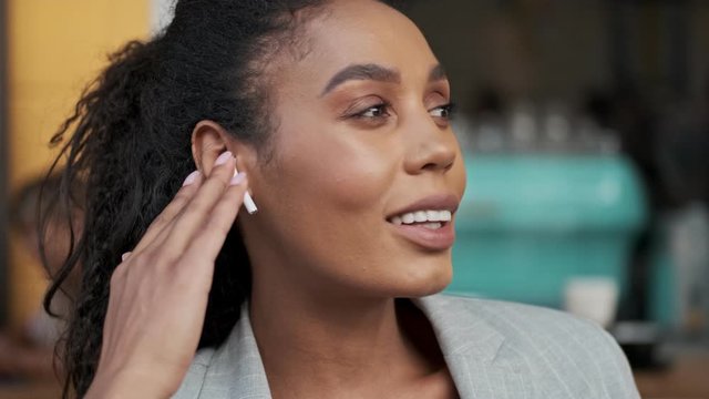 Close up view of calm young african business woman listening music with earpods while sitting outdoors in cafe