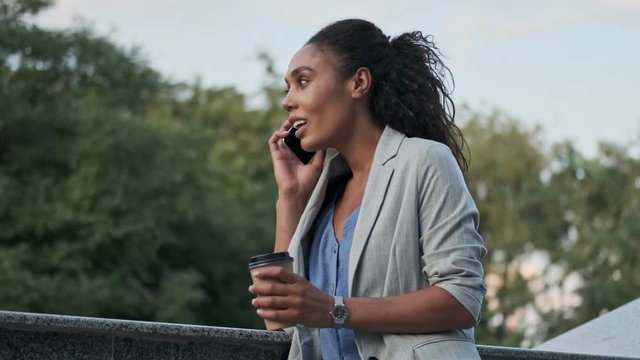 Attractive young african business woman holding takeaway coffee and talking on smartphone while resting outdoors in the park